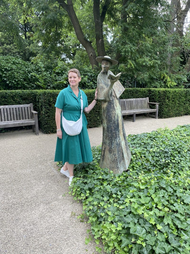 Jennifer Ellis in a teal dress beside a bronze statue of a woman reading, in a walled garden