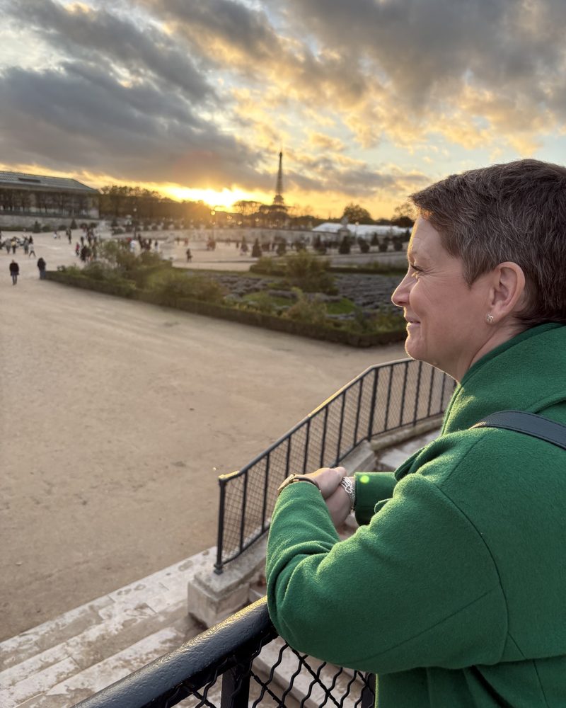 Jennifer Ellis in profile, smiling, looking out over a Paris garden at sunset in a green coat